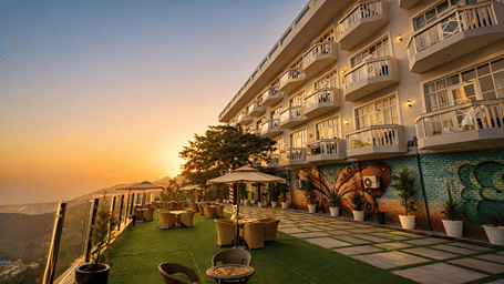 Outdoor terrace with tables, chairs, and umbrellas alongside a multi-story hotel with private balconies, overlooking a valley at sunset at DLS Highland Resort & Spa, Mussoorie.