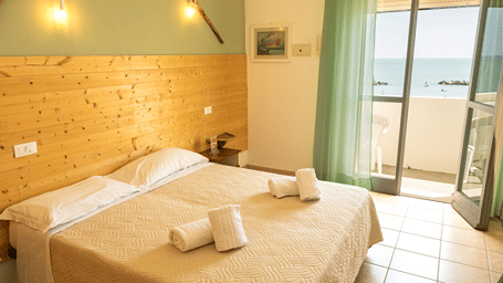 Modern resort room with twin beds dressed in crisp white linen, wooden wall paneling, and an open balcony door offering a view of the sea.