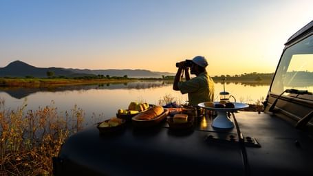 An image of a man standing near a lake with his jeep parked over looking the mountain - An image of a jeep parked amidst the rocky mountains - Utsav Camp Sariska