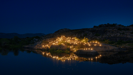 An image of an outdoor dining arrangement near a lake over viewing the mountains with bright lighting and greenery around - Utsav Camp Sariska