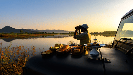 A man standing near a lake with his jeep parked over looking the mountain with binoculars near Utsav Camp Sariska.
