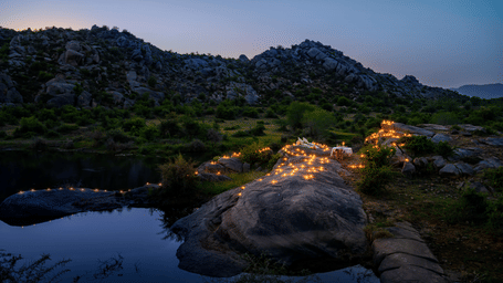 An image of an outdoor dining arrangement near a lake over viewing the mountains with bright lighting and greenery around - Utsav Camp Sariska