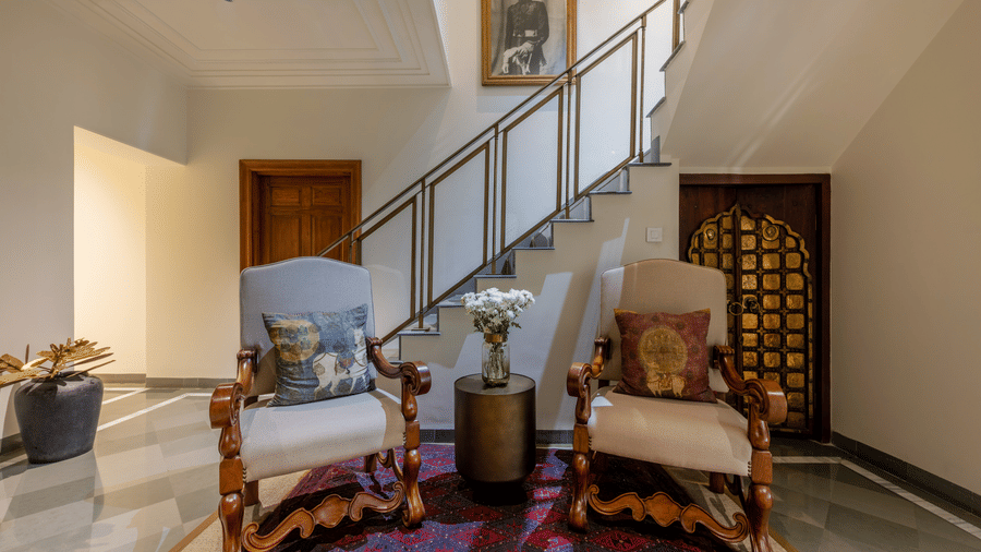 Living area beside a staircase with wooden armchairs, a patterned rug, and a wooden trunk table at Barwara Kotha, Jaipur.