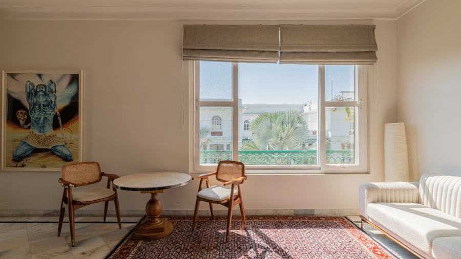 Bright living room with a sofa, cane chairs, a patterned rug, wall art, and a large window overlooking greenery at Barwara Kothi, Jaipur.