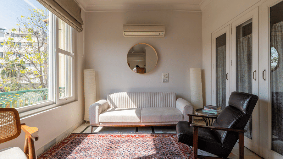 Sitting area with a sofa, leather armchair, patterned rug, round mirror, and large windows at Barwara Kothi, Jaipur.