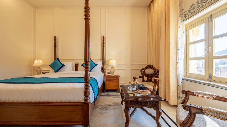 a bedroom featuring a king size bed along with two arm chairs placed in front of a coffee table with a decorative wooden frame behind it - Brij Castle, Jaipur