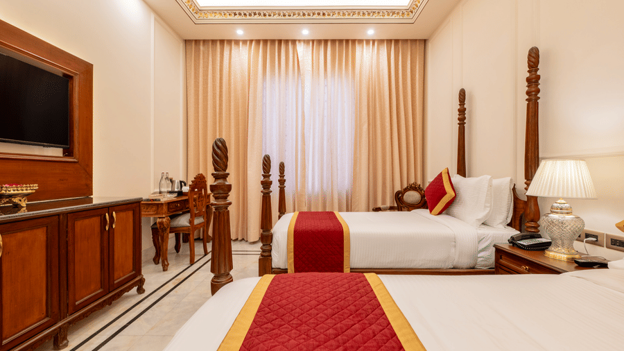 Twin poster beds with red throws, wooden console table, and wall TV in a brightly-lit bedroom at Brij Castle.