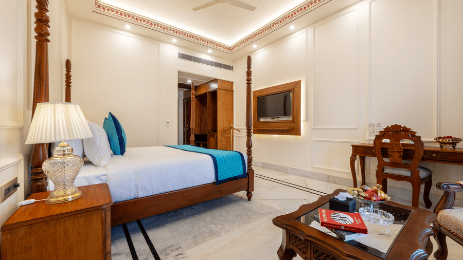 A spacious bedroom at Brij Castle featuring a poster bed with blue accents, tea table, desk, and TV.