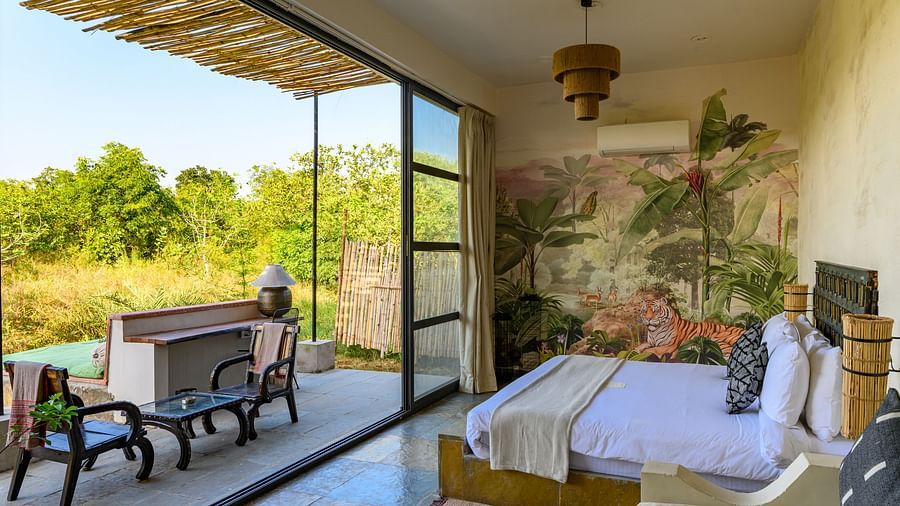 An image of a room with a queen size bed and decorative items with wall hangings and an expansive glass door with an outdoor seating arrangement - Utsav Camp Sariska