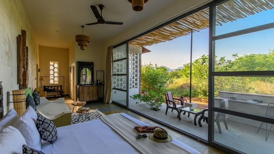 An image of a room with a queen size bed, chandelier, ceiling fan, other decorative items and an expansive glass door leading out - Utsav Camp Sariska