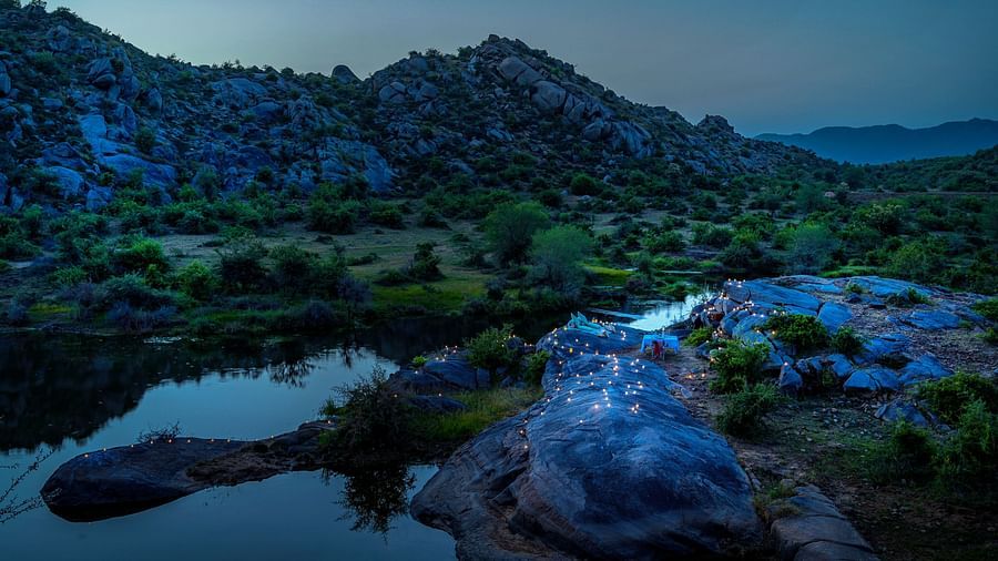 An image of an outdoor dining arrangement near a lake over viewing the mountains with bright lighting and greenery around - Utsav Camp Sariska