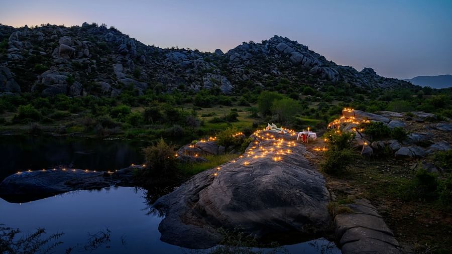 An image of an outdoor dining arrangement near a lake over viewing the mountains with bright lighting and greenery around - Utsav Camp Sariska