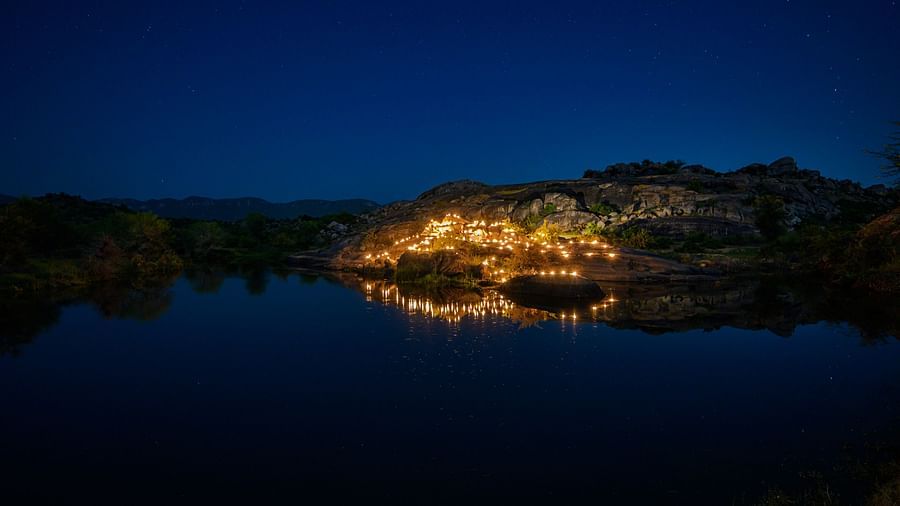 An image of a lake with rocky mountains as the background where there is a bright lit dining arrangement - An image of an outdoor dining arrangement near a lake over viewing the mountains with bright lighting and greenery around - Utsav Camp Sariska