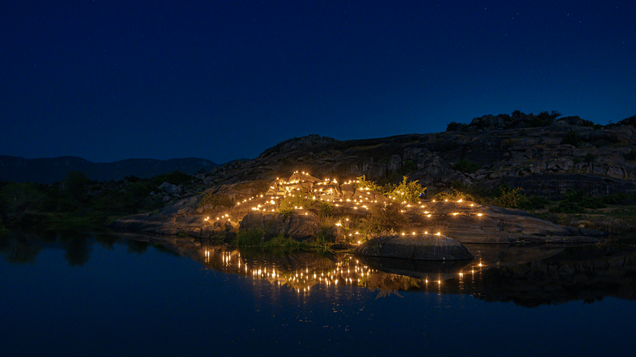 A view of an outdoor dining arrangement near a lake over viewing the mountains with bright lighting and greenery around at Utsav Camp Sariska.