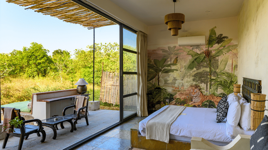 An image of a room with a queen size bed and decorative items with wall hangings and an expansive glass door with an outdoor seating arrangement - Utsav Camp Sariska