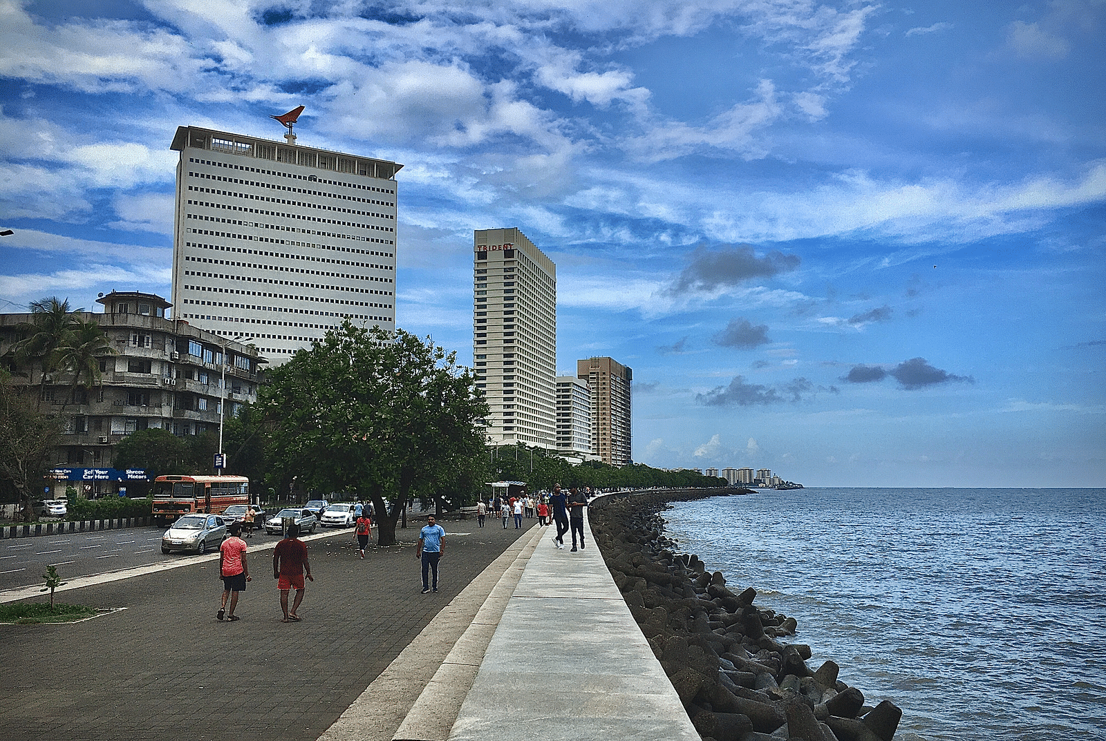 An image of a street of Mumbai near the Marine Drive with high rise buildings where are people are seen walking with a blue sky in the background