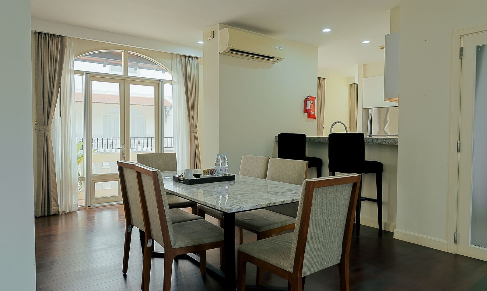 A dining room with a table and chairs, a kitchen in the background, and a balcony with curtains | Four-bedroom Suite | Central Mansions, Phnom Penh