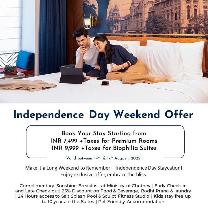 Creative hotel photography featuring a romantic couple relaxing together on a plush hotel bed while using a laptop, with Chennai's heritage architecture visible through the window, highlighting the cozy Independence Day weekend staycation experience.