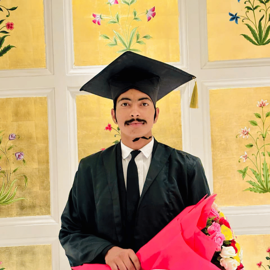 A person wearing a graduation gown and cap stands indoors. The person holds a bouquet of flowers and a box.