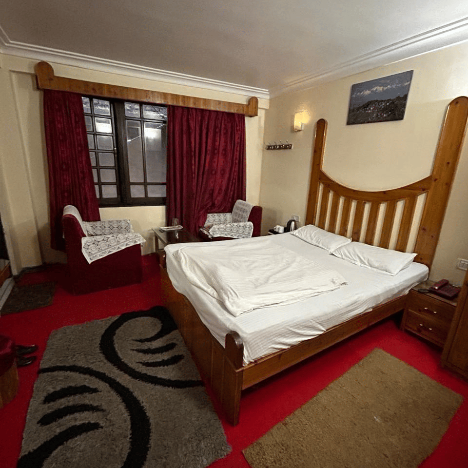 View of a bed with white linen, a wooden headboard, red carpet, and a black and white patterned rug at Hotel Dolphin Darjeeling.