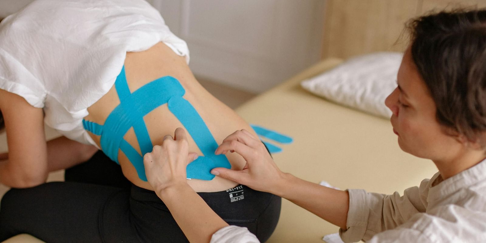  A therapist applies blue kinesiology tape to a client's lower back. The client is leaning forward on a massage table.