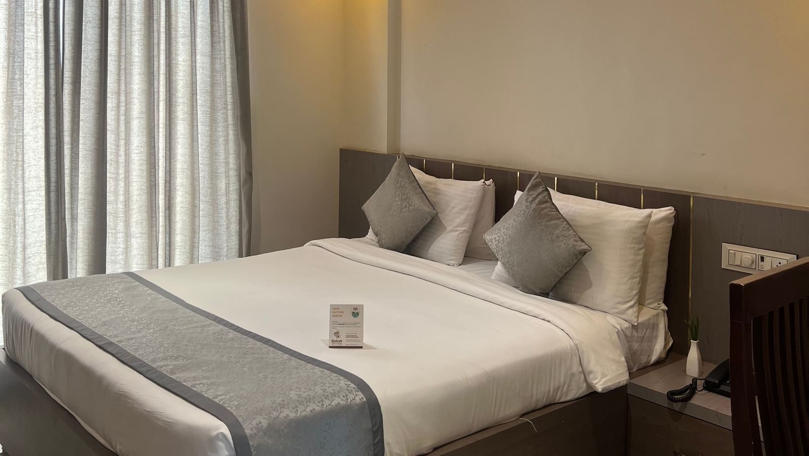 Focus view of the Daksh Bliss room at Daksh The Valley Resort, Saputara, showcasing its queen size bed, large windows with closed curtains, and warm overhead lighting.