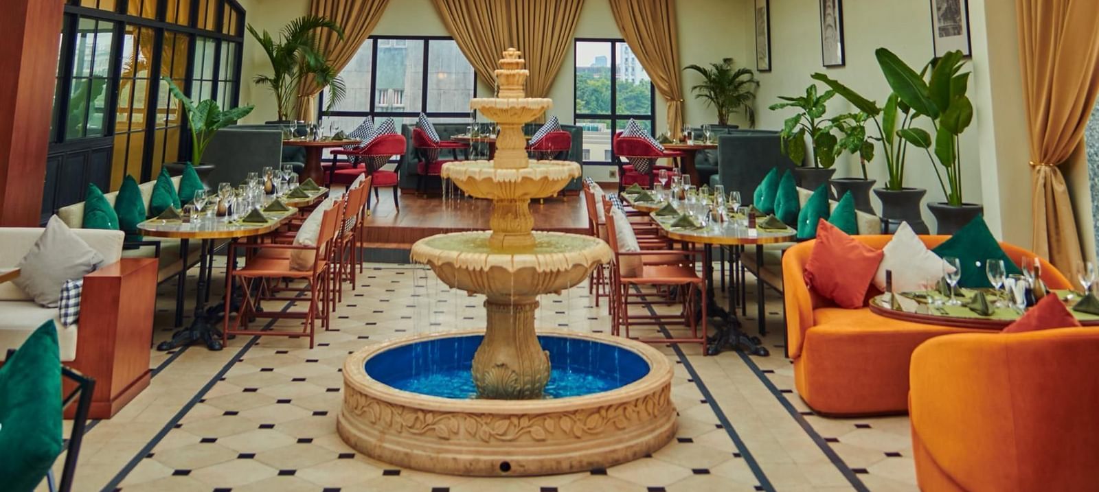 A spacious, elegant indoor Meli-Melo with a high arched glass ceiling, vibrant seating, a central fountain, chandeliers, large windows, and potted plants creating a bright, inviting atmosphere