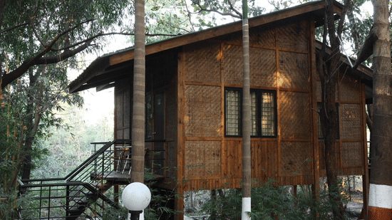 A wooden cottage with woven bamboo walls and a slanted roof, nestled among tall trees in a forested environment at Nature Trails Sajan.