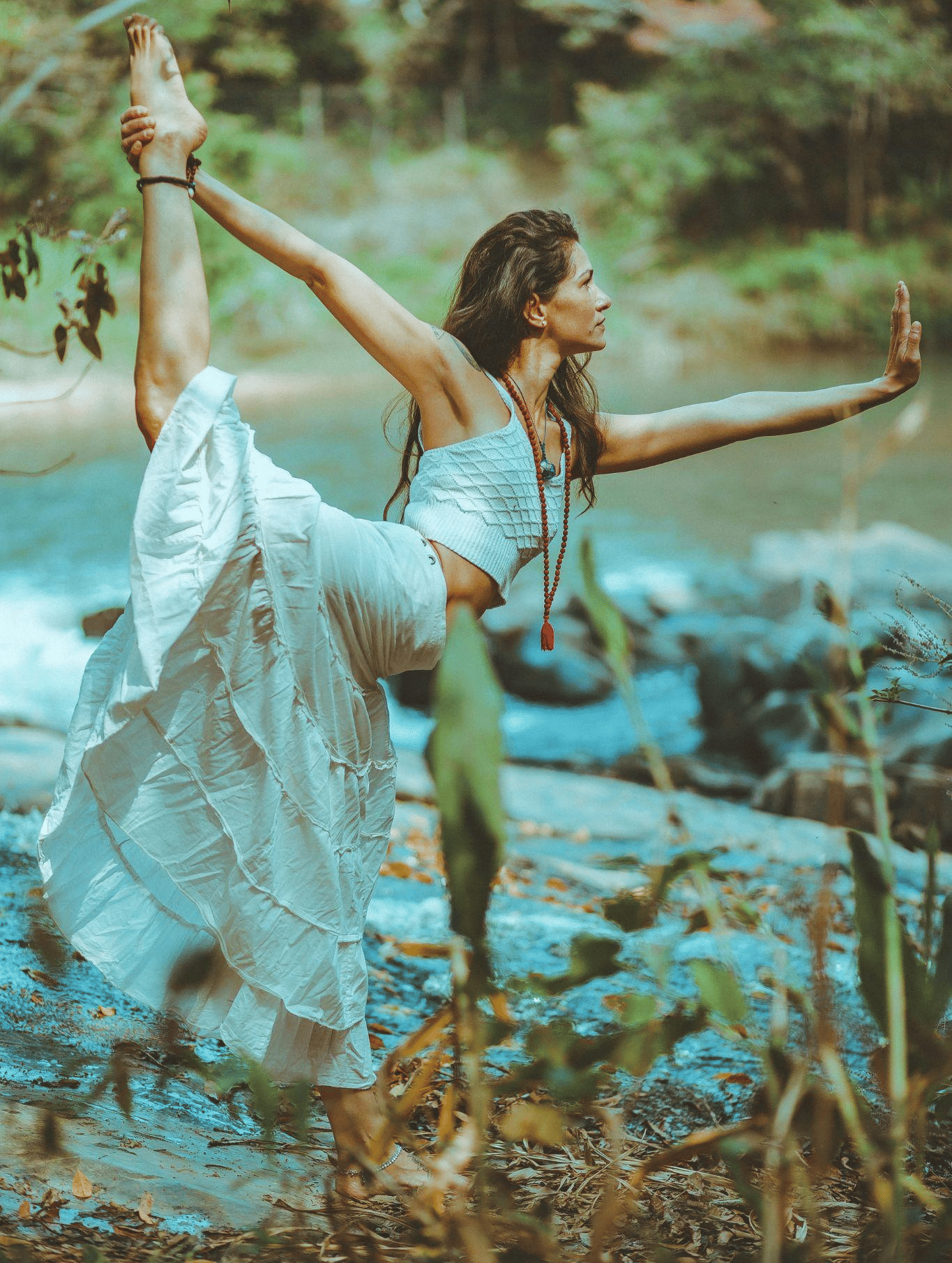 A woman in white flowy attire performs a graceful yoga pose outdoors, with a serene river and lush greenery in the background