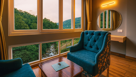 A seating area with an upholstered armchair, coffee table, large windows overlooking hills, and a wall-mounted mirror at Avianna Group of Hotels & Resorts.