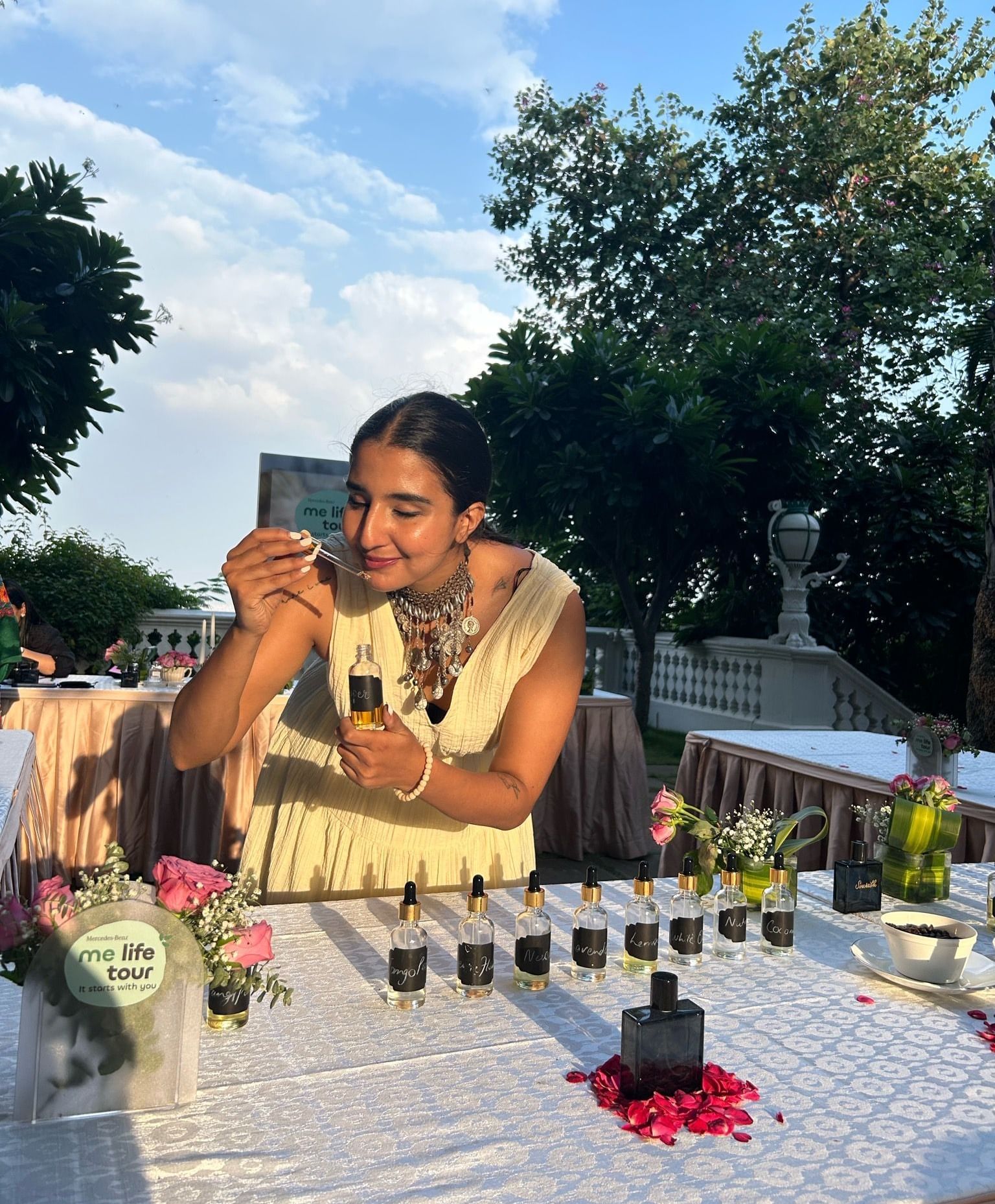 A woman in a cream dress and statement jewelry smells a scent strip while holding a perfume bottle at an outdoor fragrance table, surrounded by dropper bottles, roses, and greenery under a bright blue sky - Kastoor x Sawai Hospitality