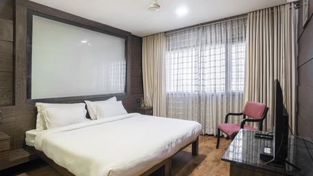 A bedroom at Infiniti Hotel, Indore featuring king size bed, television console, chair, and large window with curtains.