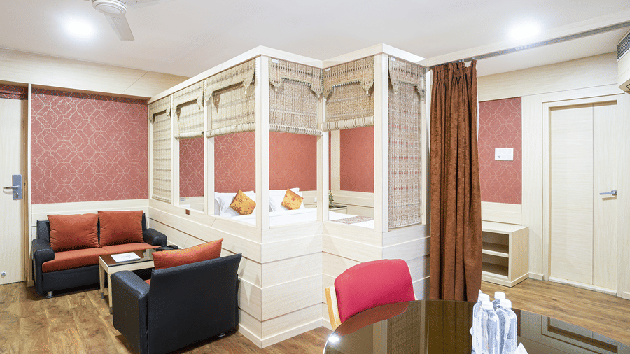 A suite with white bed, black and red sofa, wooden dining table with a pink chair and wooden partition at Infiniti Hotel, Indore.