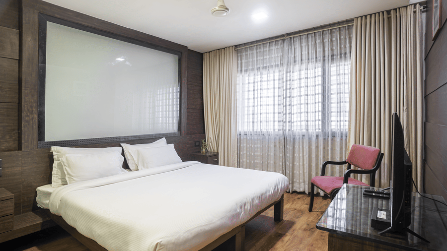 A spacious room with a king size bed, beige curtains, dark wall panelling, a pink armchair, and a T.V. at Infiniti Hotel, Indore.