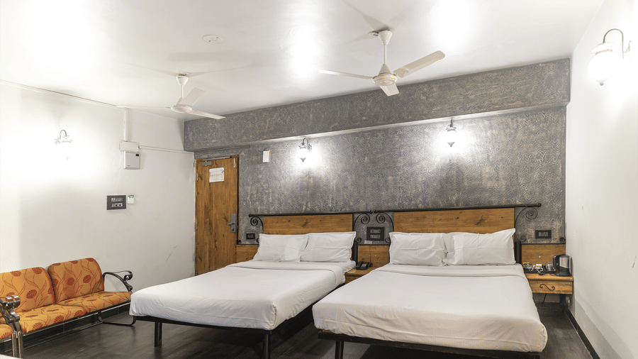 Brightly lit room featuring two double beds, a textured accent wall, wooden door, bedside tables, ceiling fans, and a small orange cushioned sofa in the corner at Infiniti Hotel, Indore.