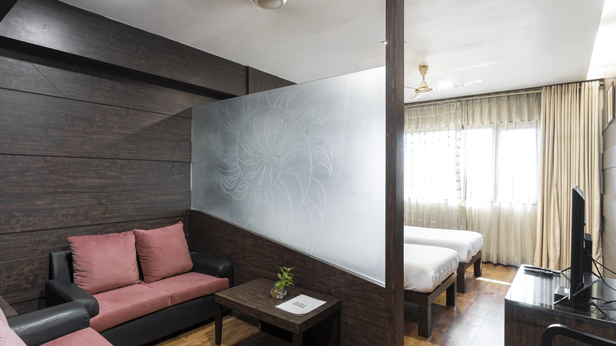 Room with sofa set, table, twin beds and large window with curtains at Infiniti Hotel, Indore.