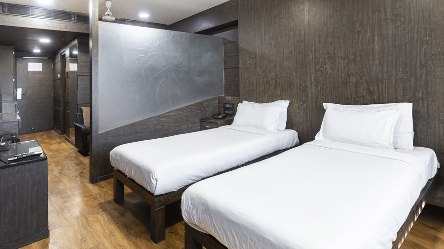 A room with two single beds, dark textured walls, a ceiling light, and a compact open wardrobe unit at Infiniti Hotel, Indore.
