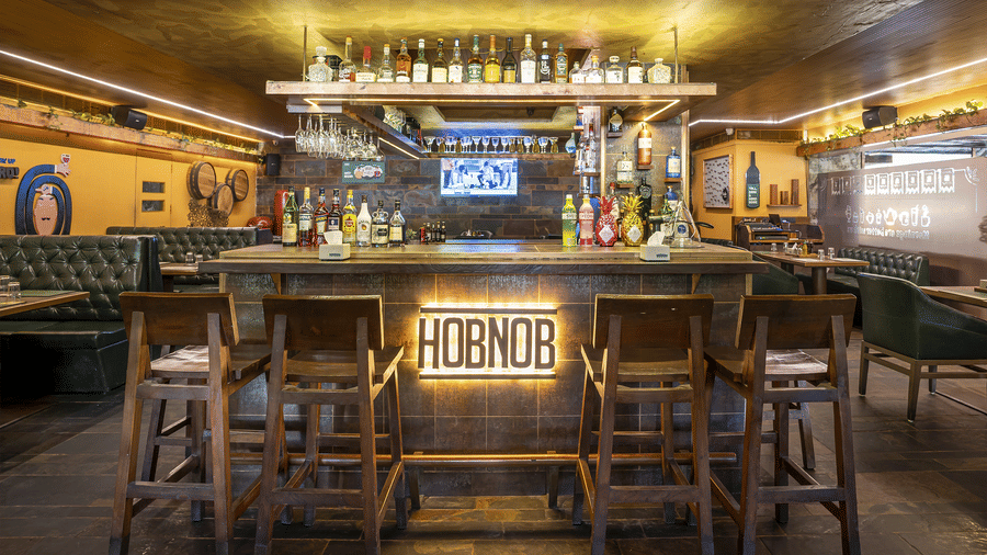 A bar counter with stools, a glowing 'HOBNOB' sign and bottles displayed behind at Infiniti Hotel, Indore.
