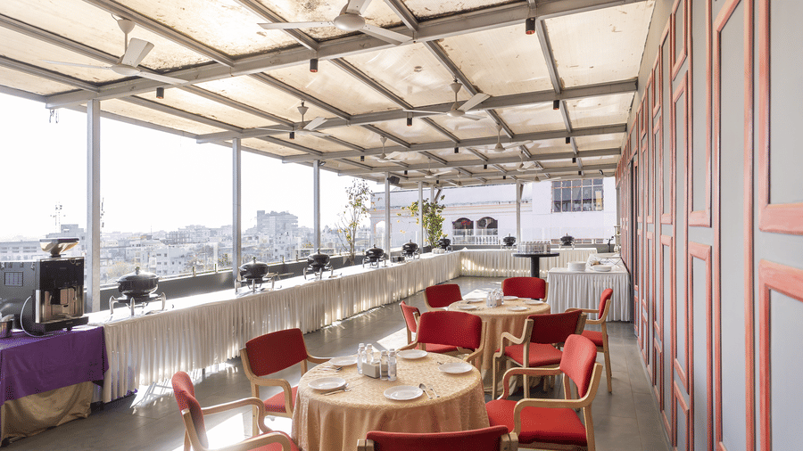 An outdoor seating area with round tables, red chairs and a roof covering with a city view at Infiniti Hotel, Indore.