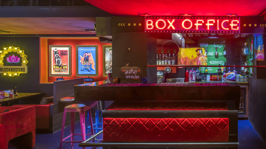 A bar counter with a glowing 'BOX OFFICE' sign, red lights and wall posters at Infiniti Hotel, Indore.