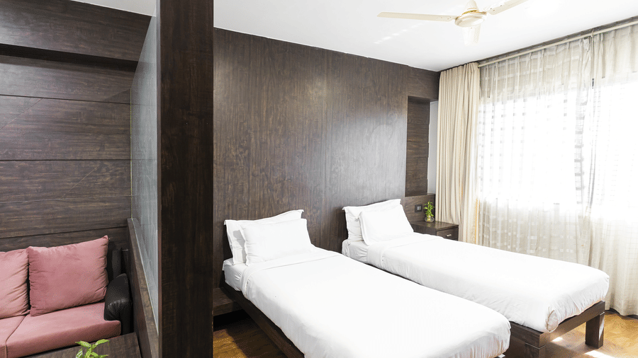 A bedroom at Infiniti Hotel, Indore featuring 2 single beds, glass room divider, sofa, and window curtains.