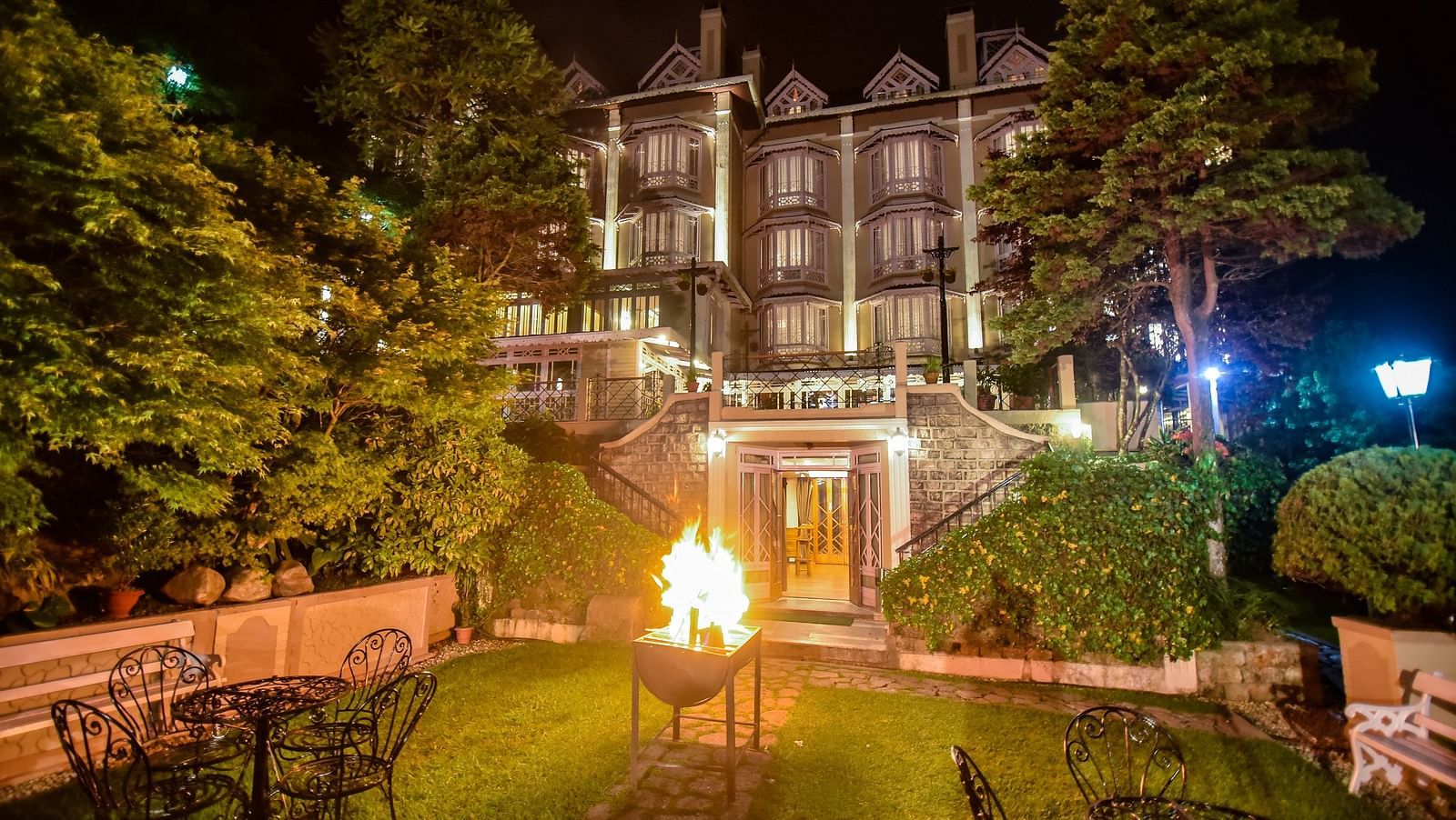 Night view of Cedar Inn, Darjeeling with a cozy outdoor fire pit, garden seating, and the warmly lit heritage building surrounded by lush greenery.
