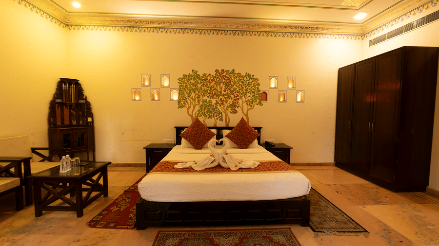 A spacious bedroom at Garh Govind with a king size bed with a tree mural on the wall behind it, a wardrobe, and seating area.