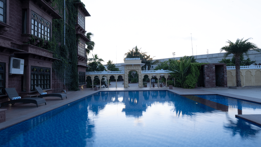 A large blue swimming pool sits next to a multi-storey building at Garh Govind, with lounge chairs on the deck and trees in the background.