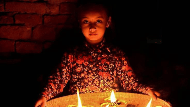 A young Nepalese child holding a clay plate filled with glowing diya oil lamps against a brick wall during Tihar in Bhaktapur, Nepal.