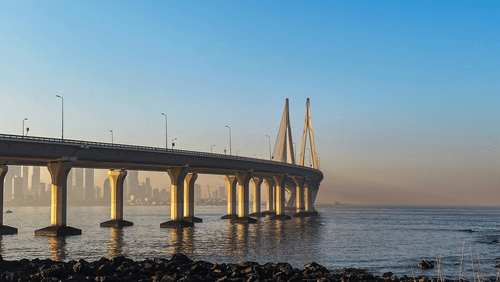 A long sea bridge stretches across calm waters, framed by a clear blue sky and rocky shoreline in the foreground.
