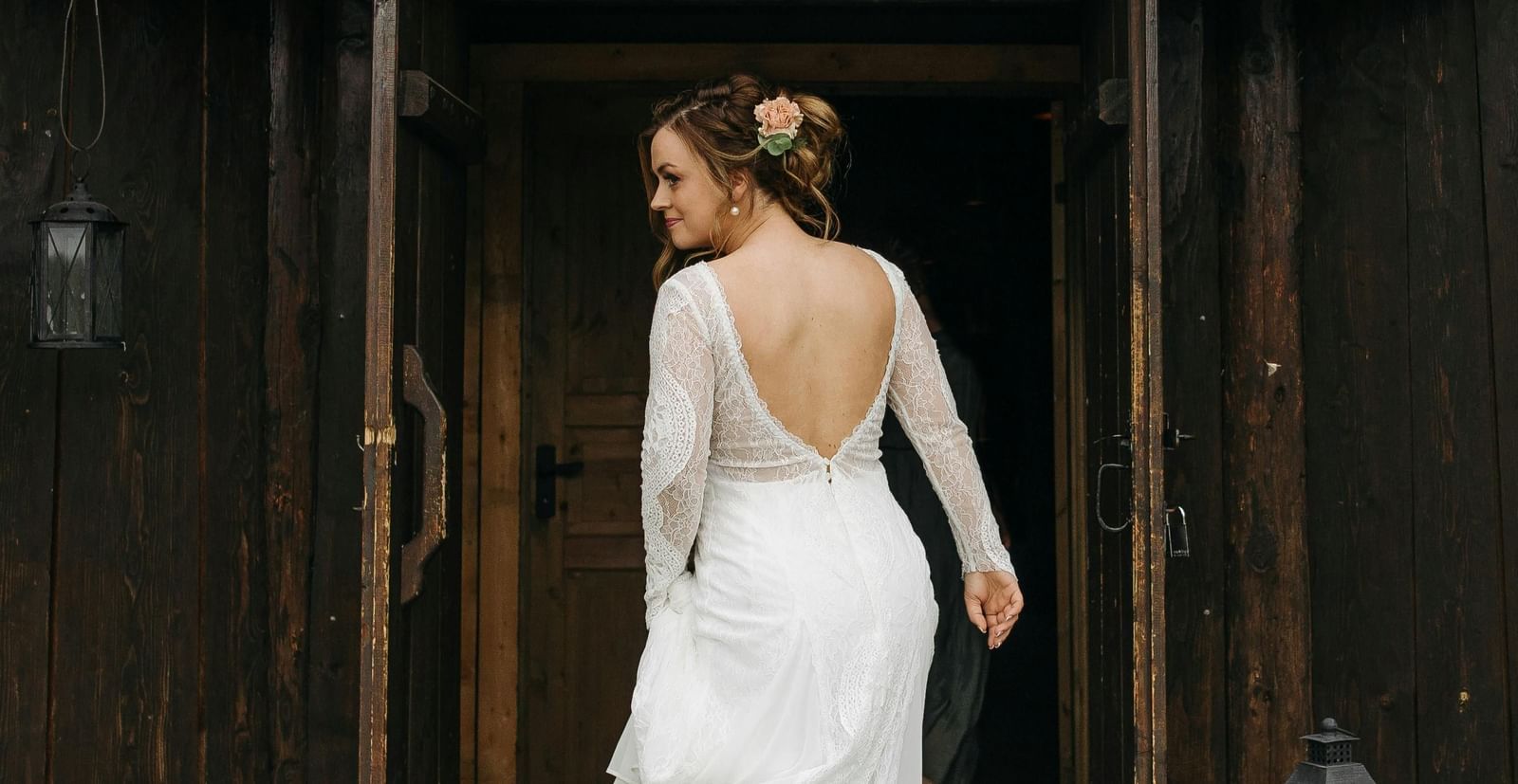 A bride in a white gown stands in a doorway of a rustic, dark-colored building, looking away