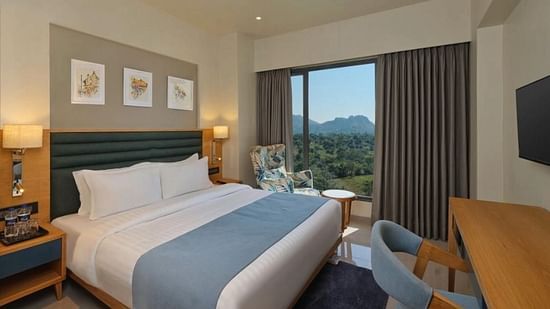 Side view of a room with a double bed and tree frames hanging on a wall and a balcony with scenic view outside an armchair and a tabe at Pride Resort Ambaji