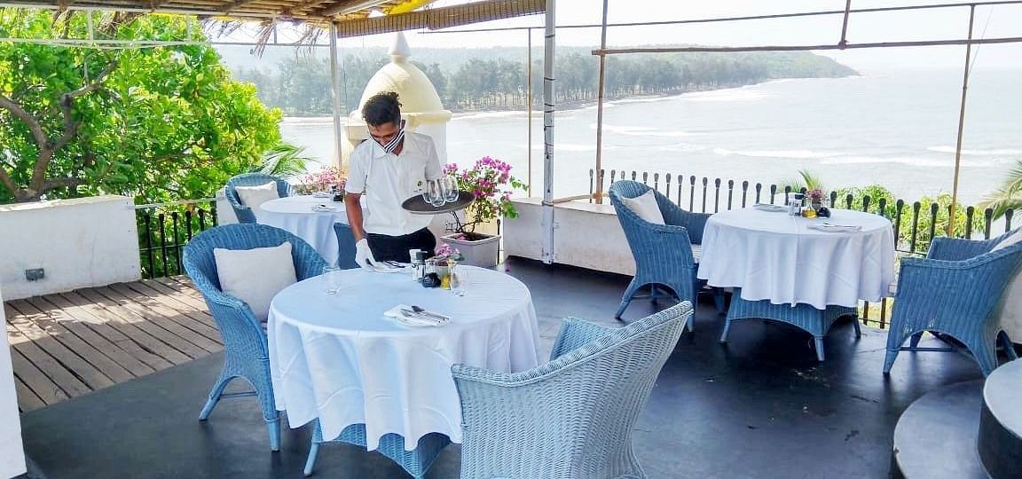 An outdoor dining area at Fort Tiracol, Goa, with tables set with white tablecloths and light blue chairs, under a shaded canopy.