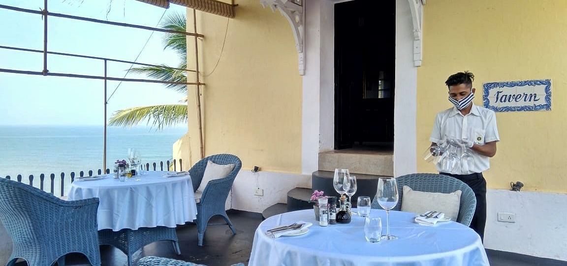 An indoor restaurant area at Fort Tiracol, Goa, with tables set with white tablecloths and wicker chairs.